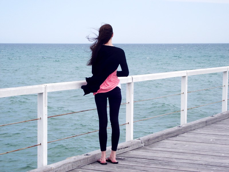 woman wearing black leggins, a pink shirt, and black cardigan looking out to the ocean from a pier. recover from an eating disorder