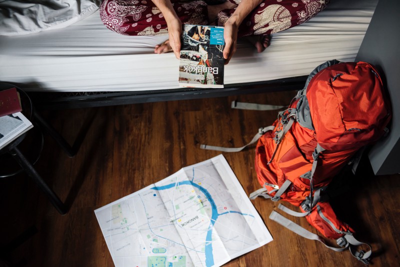 girl sitting on hostel bed holding travel guide with map and backpack on the floor in front of her