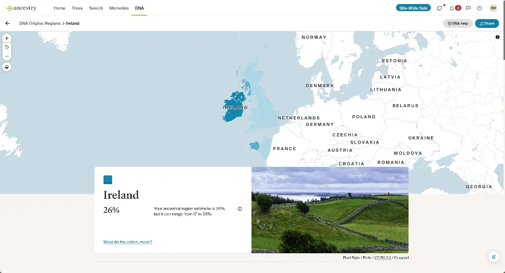 Alt text: A DNA ancestry map highlighting Ireland, shaded in blue, indicating a 26% ancestral estimate for this region, with a range from 17% to 28%. The map centers on Ireland and the surrounding areas. Below the map, a photo depicts a scenic view of the Irish countryside with green rolling hills, a stone fence, and a cloudy sky.