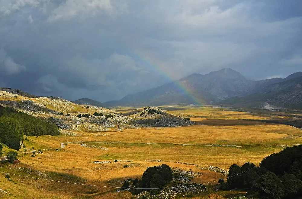 Alt text: A scenic landscape in Abruzzo, Italy, featuring golden fields stretching across a valley, surrounded by rocky hills and patches of greenery. A rainbow arches across the sky, set against a backdrop of dark clouds and distant mountains. The scene captures the dramatic beauty of the region with a mix of sunlight and shadow.