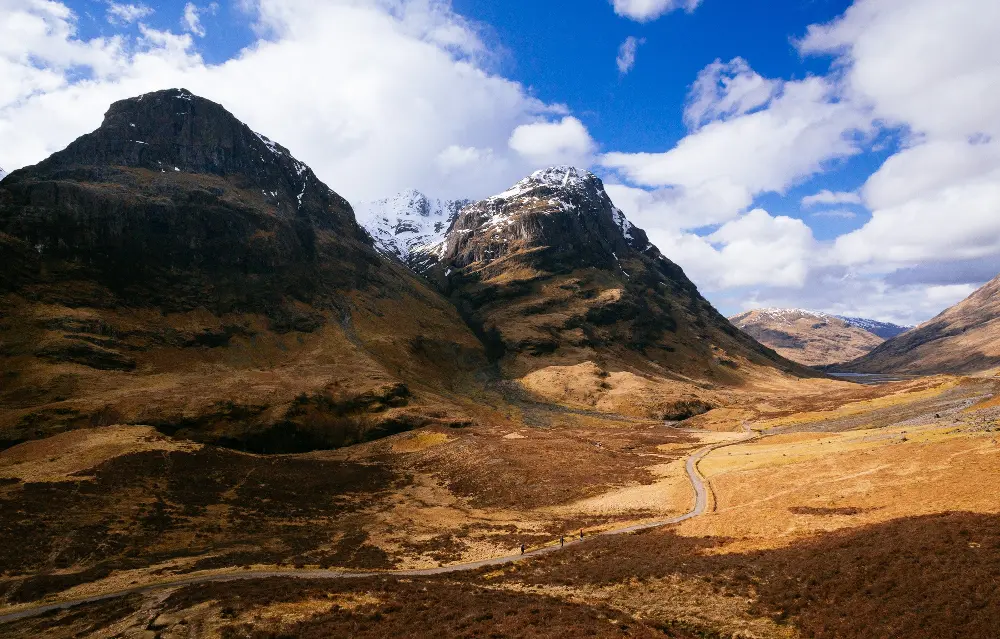 Alt text: A dramatic landscape in Glen Coe, Scotland, featuring towering, rugged mountains with patches of snow under a partly cloudy blue sky. A winding road cuts through the valley floor, surrounded by brown and golden hues of grass and heather. The scene captures the raw, natural beauty and majesty of the Scottish Highlands.