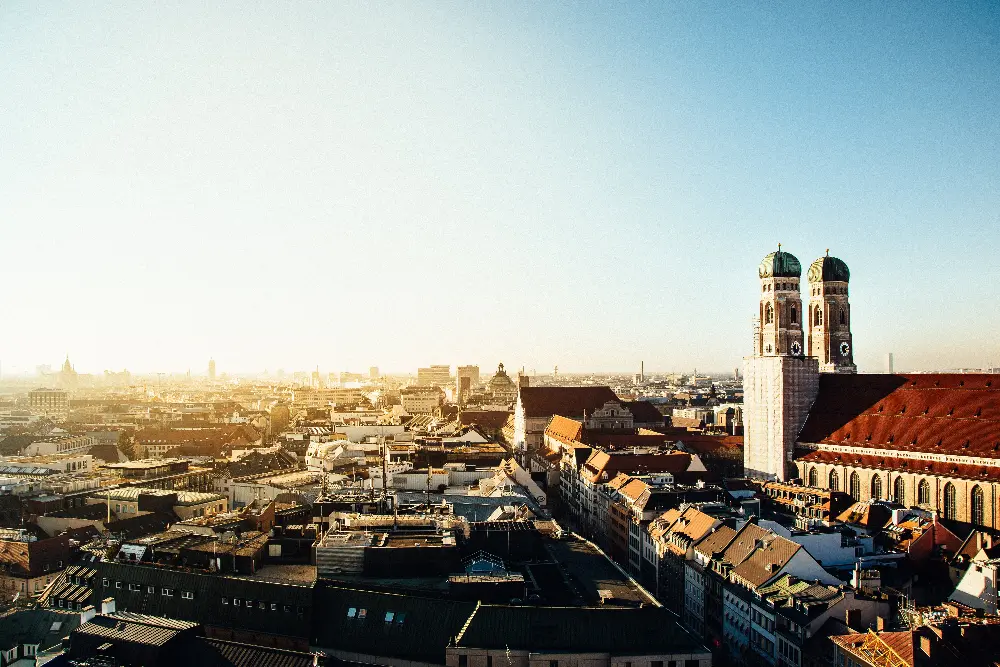 Alt text: A panoramic view of Munich, Germany, with the iconic twin domes of the Frauenkirche prominently visible on the right. The city's rooftops stretch across the frame, bathed in warm sunlight under a clear blue sky. The scene captures the historic and architectural charm of the city with its blend of traditional and modern buildings.