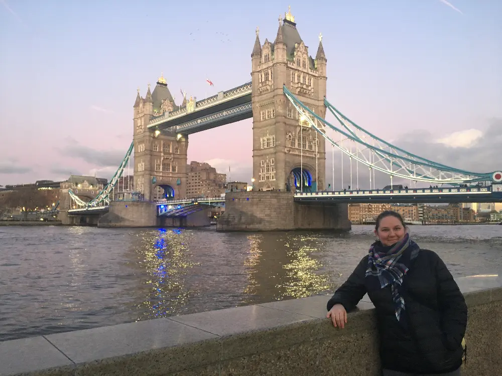 Alt text: A woman stands smiling in front of Tower Bridge in London, England, during the early evening. The iconic bridge, illuminated with blue and white lights, spans the River Thames in the background, with its twin towers rising against a pastel sky. The scene captures a peaceful moment by the river as daylight fades.