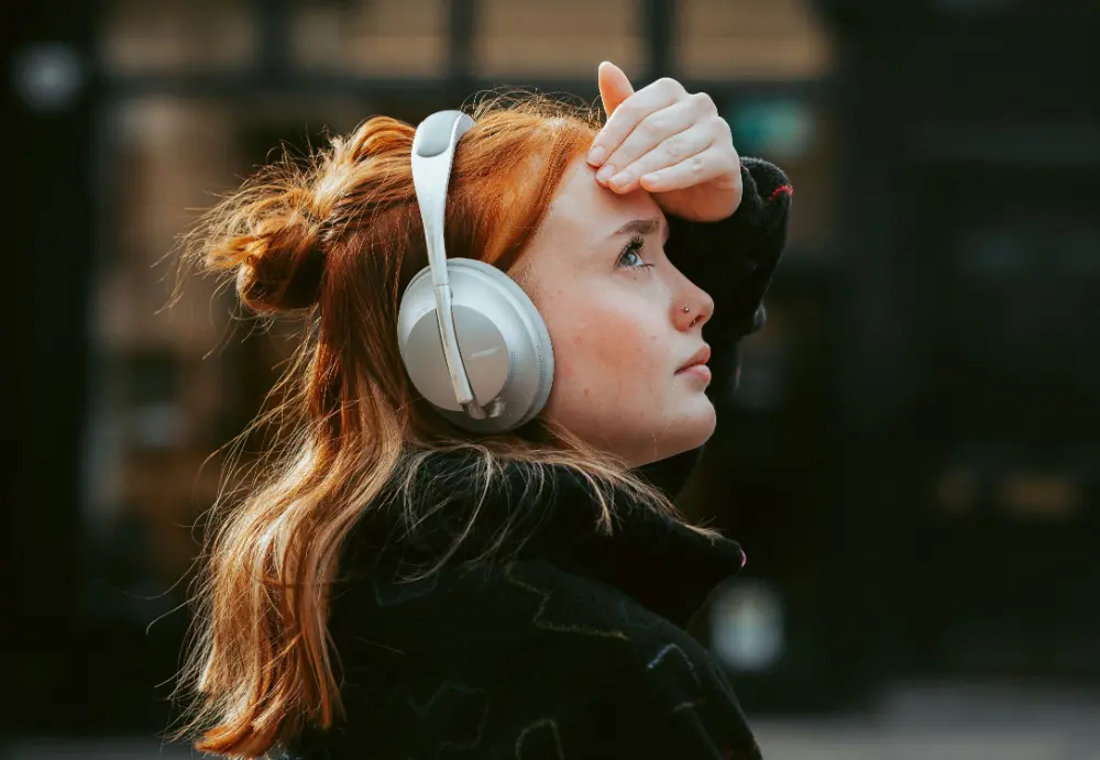 The image shows a young woman with red hair wearing over-ear headphones, gazing upward while shielding her eyes with her hand. The background is blurred, suggesting an outdoor or urban setting.