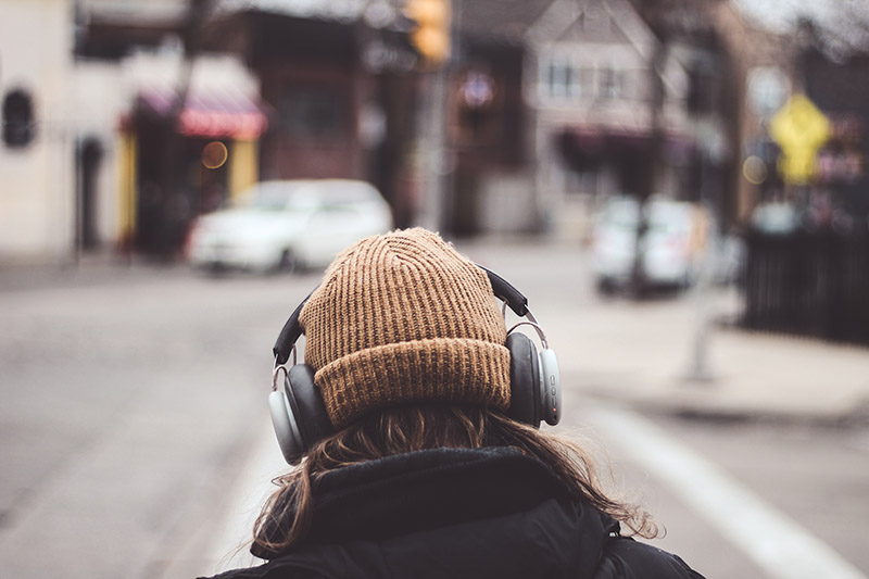 A person from behind wearing a beige knit hat and headphones, looking out onto a blurred city street scene.