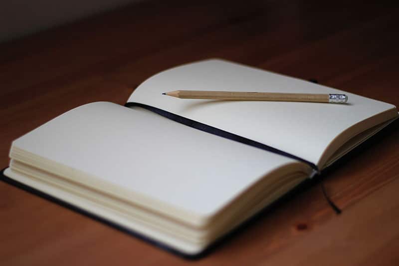 An open blank notebook lies on a wooden surface, accompanied by a sharpened pencil resting on its right page, evoking a sense of readiness for writing or drawing.