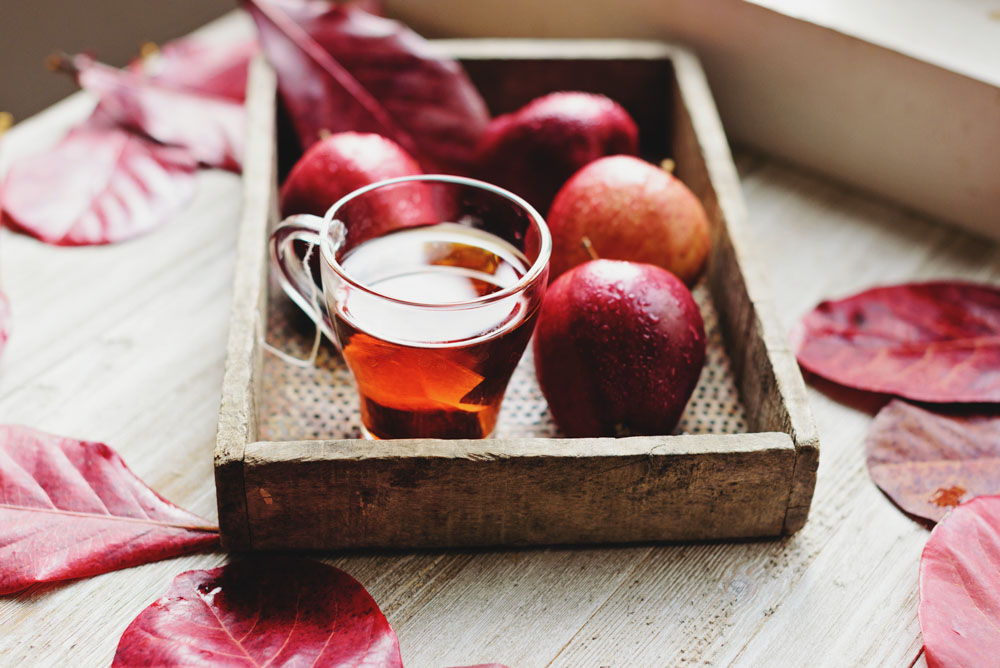 A wooden tray holding a glass mug of hot apple cider and red apples with red leaves spread out around it on a wooden table. fall hygge songs