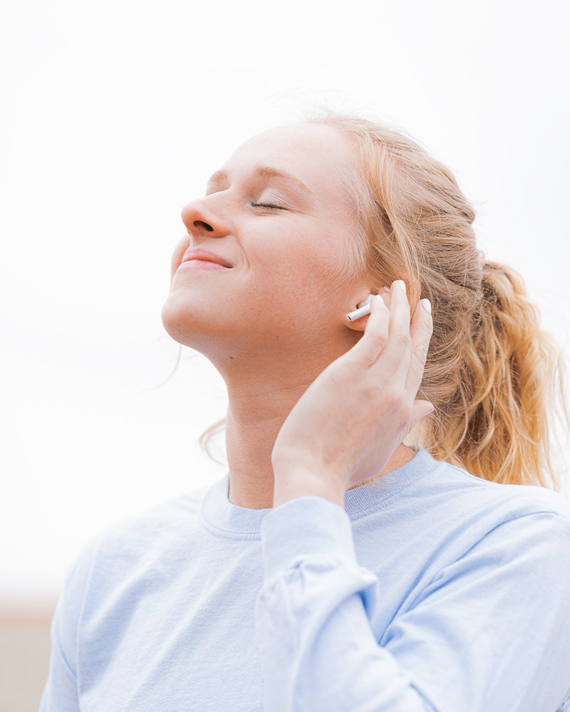 A young girl with long blonde hair in a ponytail, a long-sleeved lavender-colored shirt, and closed eyes listening to music using AirPods. spring hygge songs