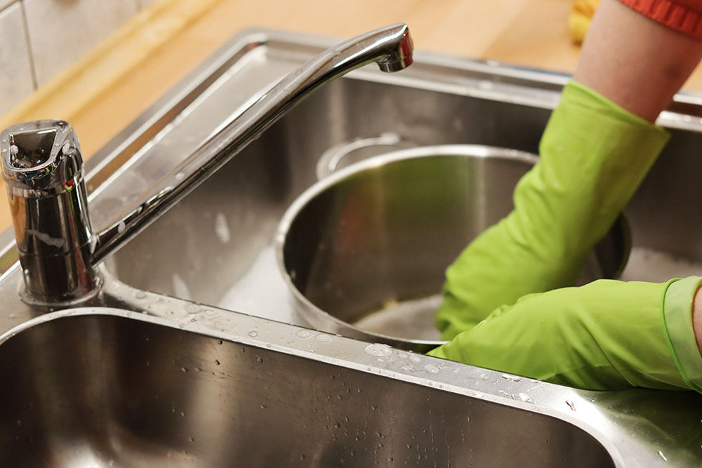 A person wearing green gloves washing a large pot in a sink.