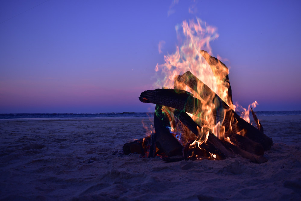 Close up of a bonfire on a beach during a purple and pink sunset. summer hygge songs