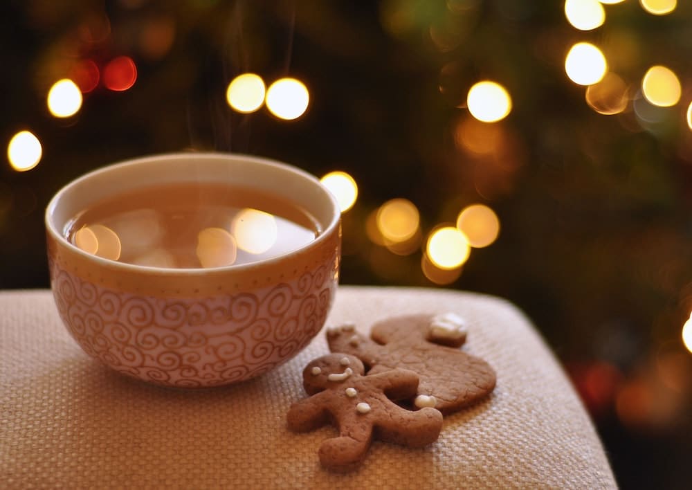 a cup of tea and gingerbread cookies in front of the blurry lights of a Christmas tree. in a blog post about cozy Christmas songs.