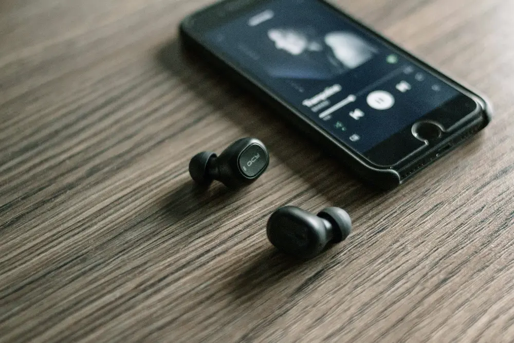 Wireless earbuds resting next to a smartphone displaying a music player interface with an artist's photo on the screen, all on a wooden surface.