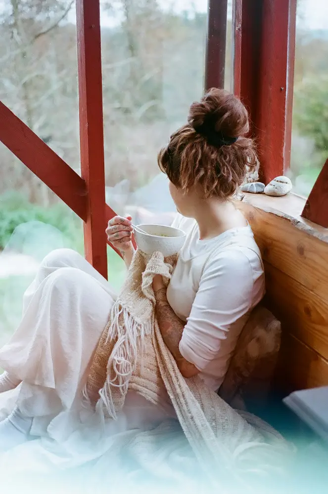 A serene moment captured of a woman sitting cozily by a cabin window, wrapped in a cream knit blanket, gently holding a large ceramic mug, with a soft focus on the lush greenery outside.