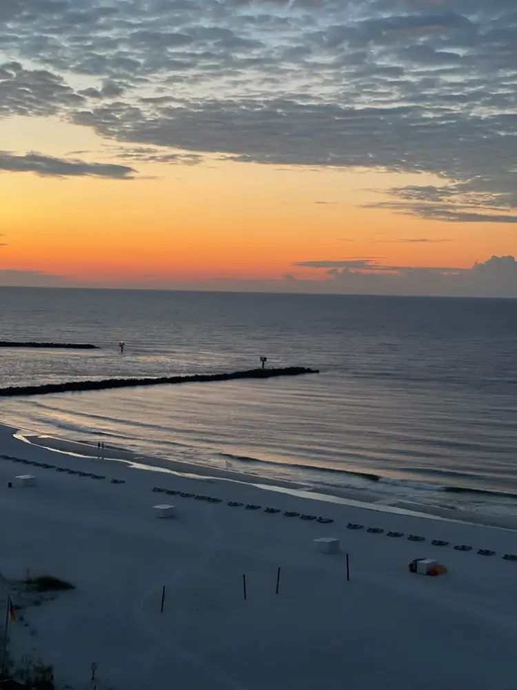 A serene beach scene at sunset, with the sky painted in shades of orange and pink, gradually fading into twilight. The calm ocean waves lap against the sandy shore, lined with empty lounge chairs, while a jetty extends into the water, adding to the tranquil atmosphere.