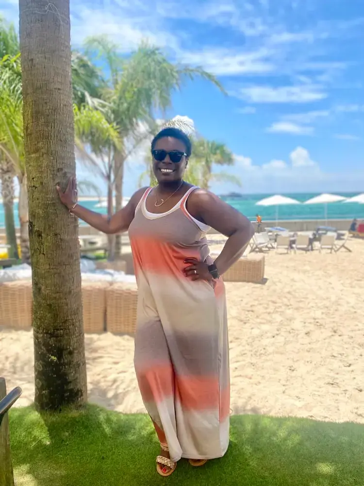 A woman in a sleeveless, gradient dress stands confidently next to a palm tree on a sunny beach. She wears sunglasses and smiles, with a backdrop of sandy shores, lounge chairs, and the ocean under a clear blue sky.