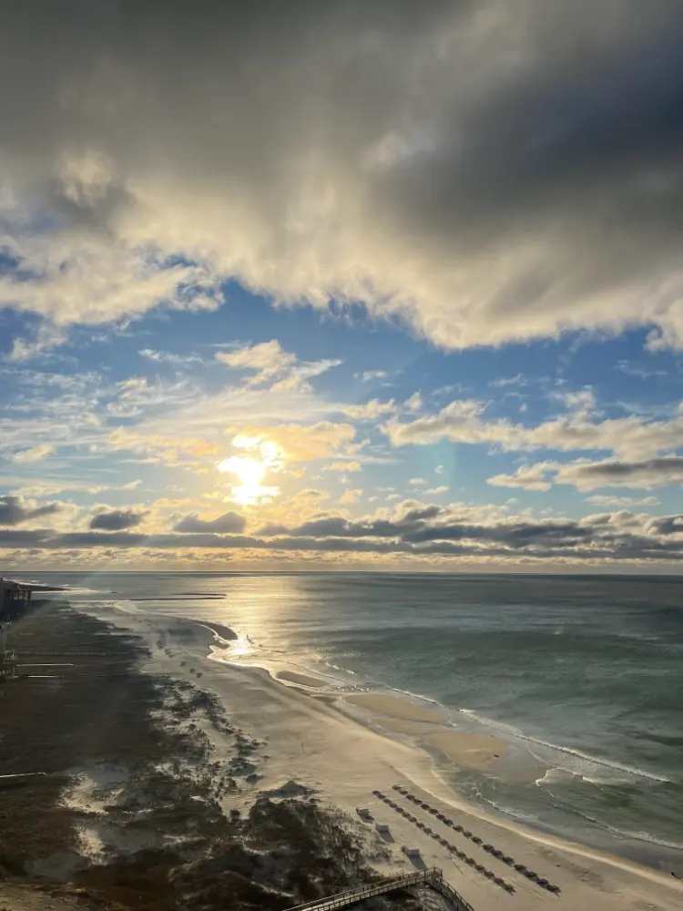 A dramatic beach scene captured at sunrise, with the sun rising over the horizon, casting a golden glow on the ocean and sandy shore. The sky is filled with a mix of bright clouds and darker storm clouds, creating a striking contrast. The beach is quiet and empty, enhancing the peaceful ambiance.