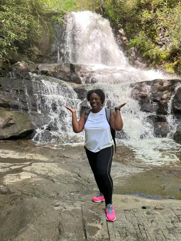 A woman in athletic wear and bright pink sneakers smiles and gestures with her hands while standing in front of a cascading waterfall in a lush, forested area. The scene exudes a sense of adventure and joy amidst nature.