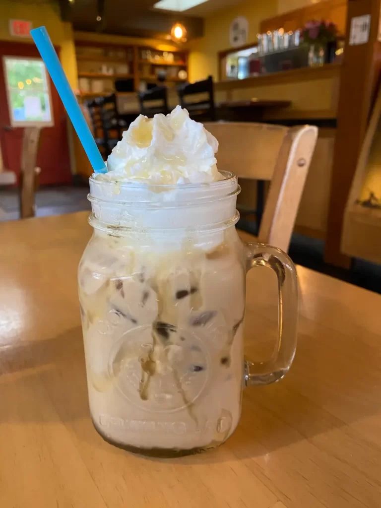 A creamy iced coffee served in a mason jar with a handle, topped with a swirl of whipped cream and drizzled with honey. A bright blue straw sticks out of the drink, contrasting with the neutral tones. The background features a cozy café setting with wooden tables, chairs, and warm lighting.
