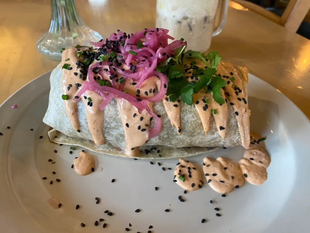 A generously sized burrito on a white plate, drizzled with a creamy orange sauce and topped with pickled red onions, black sesame seeds, fresh cilantro, and chopped chives. Some of the sauce has dripped onto the plate, adding to the presentation. In the slightly blurred background, a frothy iced coffee sits on the wooden table, along with a glass vase.