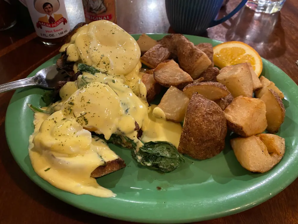This image shows a hearty breakfast plate featuring Eggs Benedict topped with rich hollandaise sauce over spinach and mushrooms on toasted bread. It's served with a generous portion of crispy roasted potatoes and a slice of orange for garnish. A coffee mug and hot sauce bottles are visible in the background, hinting at a cozy diner setting.