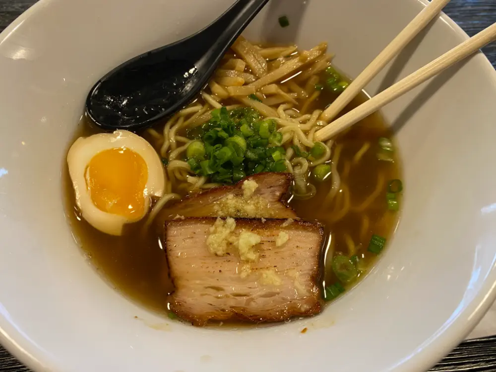 A bowl of shoyu ramen with a clear, golden-brown broth, topped with a slice of tender braised pork belly garnished with grated ginger, a soft-boiled egg with a slightly runny yolk, chopped green onions, and bean sprouts. Wooden chopsticks rest on the side, lifting some of the curly noodles, while a black spoon sits in the broth. The dish is served in a white bowl on a dark wooden table.