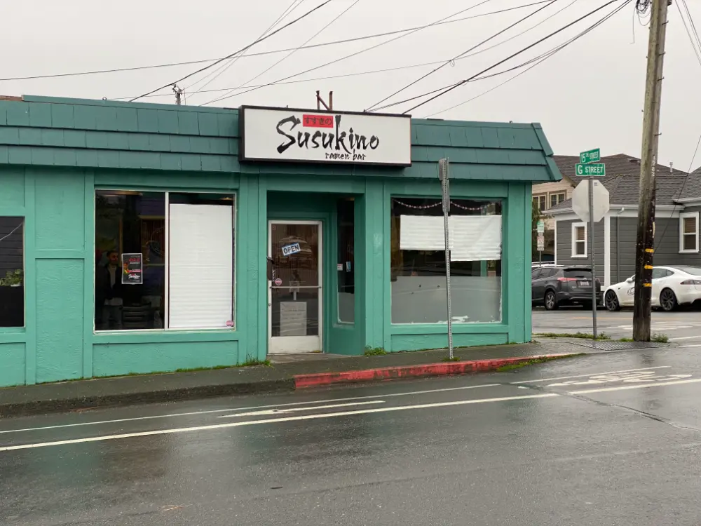 A small ramen bar named 'Susukino' with a teal exterior and a black and white sign above the entrance. A small 'OPEN' sign hangs on the glass door. The building sits on a rainy street corner, with wet pavement reflecting the overcast sky, and a few parked cars in the background.