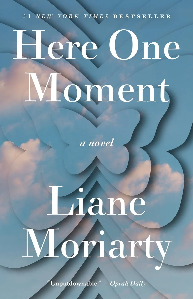 Book cover for "Here One Moment" by Liane Moriarty featuring layered blue shapes resembling a butterfly over a soft cloudy sky background. The title appears in large white serif text with "a novel" beneath it and a small line at the top reading "A New York Times Bestseller."